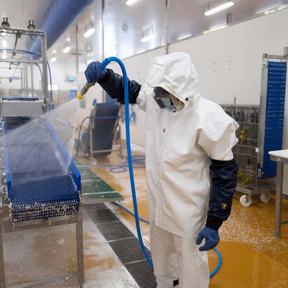 Person in a white protective suit and mask using a spray gun in a factory, setting, wearing Guy Cotten Heracles Jacket and Bib & Braces and Maree Aqua Cuffs.