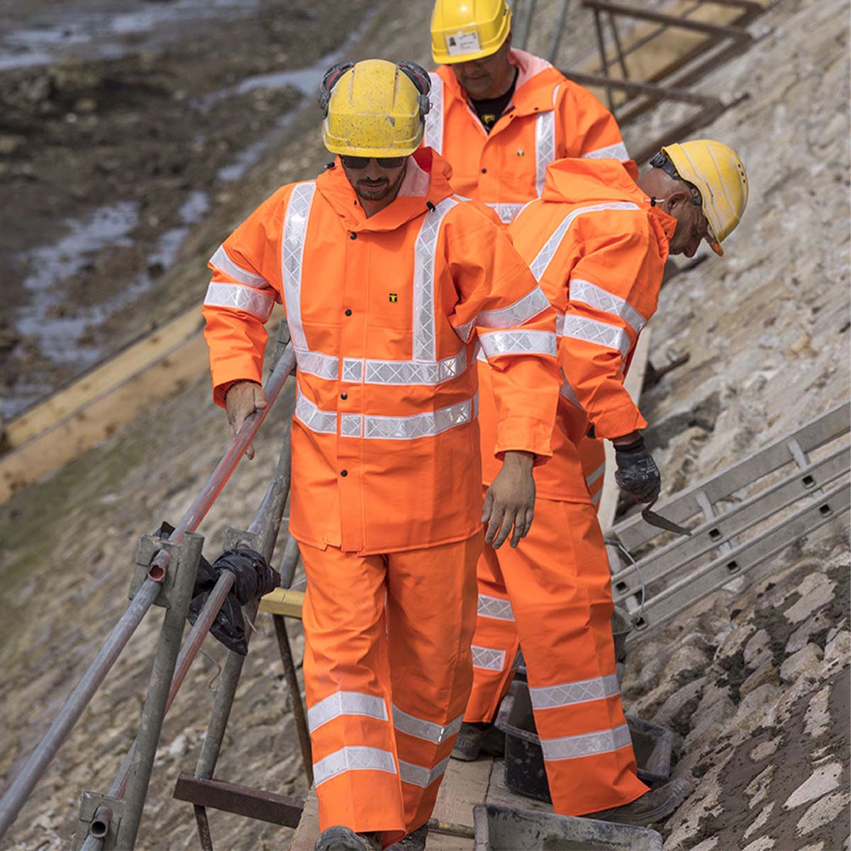 Three construction workers in orange uniforms and yellow helmets on a construction site, wearing Guy Cotten Isoflash waterproof jacket and Poulflash waterproof trousers.