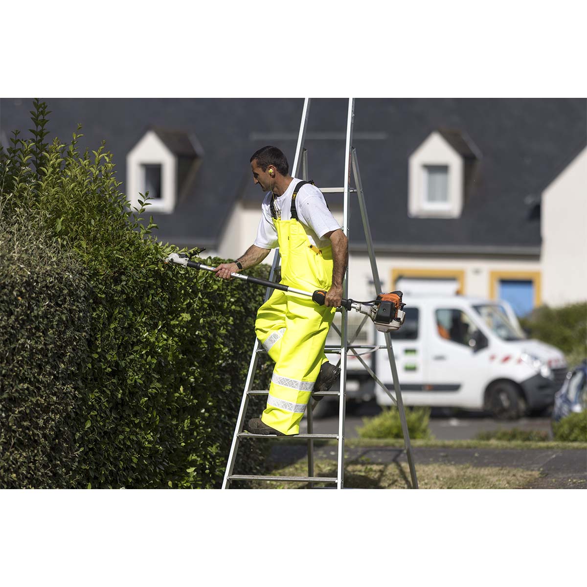 Person in Guy Cotten Hitflash high-visibility waterproof safety gear using a hedge trimmer on a ladder.